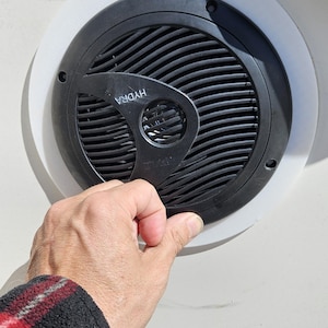 Puede incluir: Un altavoz marino circular negro con una rejilla de patrón ondulado y la palabra HYDRA impresa en blanco. El altavoz está montado dentro de un anillo adaptador circular de plástico blanco, instalado contra una superficie plana color hueso.