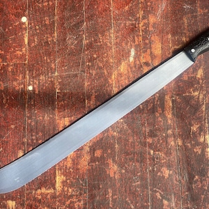 May include: A silver machete with a black rubber handle. The machete is lying on a wooden surface.