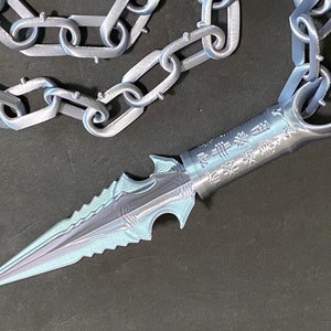 Może przedstawiać: Srebrny metalowy sztylet z przymocowaną łańcuchem. Sztylet ma ostry czubek i ząbkowane ostrze. Rączka jest grawerowana japońskimi znakami.