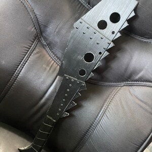 以下が含まれることがあります： ギザギザの刃とテクスチャ加工されたハンドルが付いた黒いプラスチック製の小道具武器。武器にはリベットと穴のデザインが施されています。