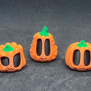 May include: Three orange pumpkin-shaped figurines with green stems and black eyes. The pumpkins have a smiling face and are made of a smooth, matte material.