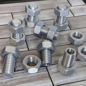 Könnte beinhalten: Eine Sammlung von silbernen Muttern und Schrauben aus Metall, einige sind zusammengesetzt, andere sind getrennt. Die Muttern und Schrauben liegen auf einer Holzoberfläche.