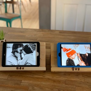 May include: Two wooden tablet stands with personalized names "mav" and "gus" engraved on the front. Each stand holds a tablet with a different photo displayed on the screen.