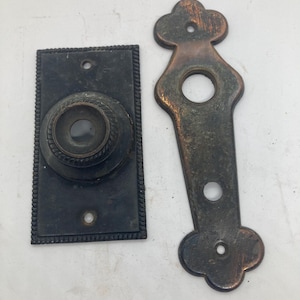 May include: Two antique door hardware pieces. One is a rectangular plate with a circular knob and rope-like trim. The other is a decorative, elongated plate with a clover-like top and bottom. Both pieces have a dark, aged finish.