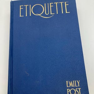 May include: A hardcover book titled "ETIQUETTE" in gold lettering on a dark blue cover. The author's name, "EMILY POST", is printed in gold at the bottom right corner. The book appears to be in good condition.