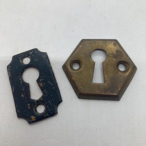 May include: Two metal keyhole escutcheon plates. One plate is rectangular with rounded edges and a distressed black finish. The second plate is hexagonal with a tarnished brass finish. Both plates feature a central keyhole and two screw holes.