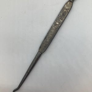 May include: A vintage metal button hook with a loop at one end and a curved hook at the other. The text "QUEEN QUALITY SHOES" is etched on the handle. The tool is a dark, aged silver color.