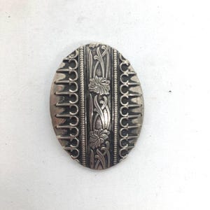 May include: An oval-shaped silver-toned metal brooch featuring a central vertical panel with embossed floral and vine motifs, flanked by two decorative borders with repeating arched patterns and a beaded edge detail on a metallic surface.