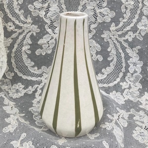 May include: A white ceramic vase with vertical olive green stripes. The vase has a flared shape and a narrow neck. The stripes are evenly spaced and run from the base to the top of the vase. The vase is a decorative item.