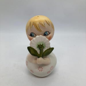 May include: A vintage ceramic figurine of a child holding a white daisy. The figurine has a blonde head, blue eyes, and a white dress with pink floral accents. The flower has green leaves and a white petal design.