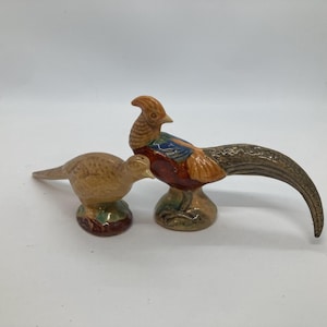 May include: Two ceramic pheasant figurines. The smaller bird is light brown with a textured surface. The larger bird features a tan head, a dark red chest, blue wing accents, and a long, curved, mottled brown tail, both standing on bases.