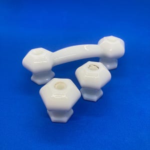 May include: A set of three white ceramic cabinet hardware pieces consisting of one arched handle and two matching hexagonal knobs. Each piece features a smooth, glossy finish with a central mounting hole, designed for furniture or drawer attachment.