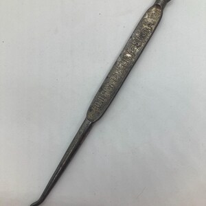 May include: An antique metal button hook with the text "QUEEN QUALITY SHOES" embossed on the handle. The hook has a loop at the top and a curved hook at the bottom, with a dark, aged finish.