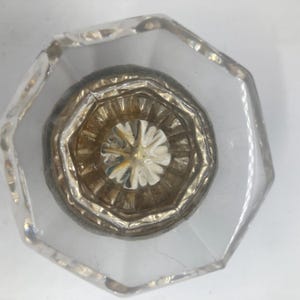 May include: A clear glass knob with an octagonal shape and a central metallic insert. The center features a circular, fluted design with a reflective gold-toned finish and a star-like pattern radiating from the middle point of the hardware.