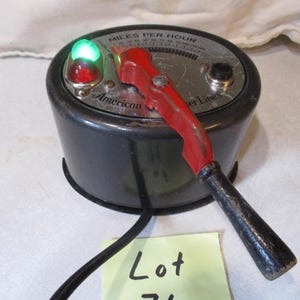 May include: Vintage black and silver speedometer with a red handle and a green light. The gauge reads "Miles Per Hour" and has the text "American Flyer Lines". A black cord and a yellow sticky note with the text "Lot 71" are also visible.