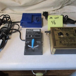 May include: Two electric train transformers, one blue and one gold, with a black power cord and plug. The blue transformer is labeled "Tyco" and the gold transformer is labeled "Trainpack". A yellow sticky note with the text "HO Lot 46" is visible in the background.