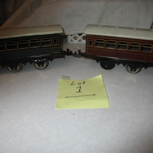 May include: Two vintage toy train cars. One is dark green, the other is maroon, both with cream-colored roofs. The cars have multiple windows and are connected. A yellow sticky note with "Lot 1" is in the foreground.