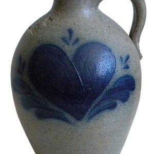 May include: A stoneware jug with a handle and a dark blue heart design. The jug has a light gray color and a textured surface. The heart is surrounded by blue leaf-like accents. The jug has a small opening at the top.