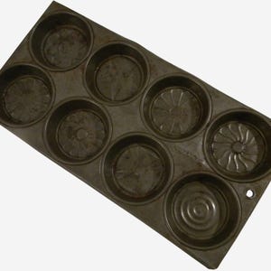 May include: An antique, dark-colored muffin tin with eight circular molds. Each mold has a decorative pattern at the bottom. The tin is rectangular with a small hole on one end, suggesting it may be hung for storage. Baking and kitchenware.