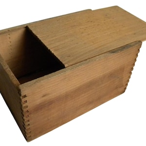 May include: A rectangular wooden box with a sliding lid, crafted from light-colored wood. The box features dovetail joints at the corners and a partially open lid, revealing the interior. The wood grain is visible, giving it a rustic appearance.