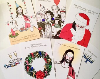 May include: A selection of humorous greeting cards with festive themes. The cards display illustrations of Jesus, Santa Claus, and other holiday imagery, accompanied by witty text including "Happy Hanukkah" and "Have yourself a very Agnostic Christmas."