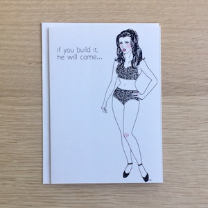 May include: A white greeting card with the text "If you build it, he will come..." features a black and white illustration of a woman in lingerie. The woman is drawn with black lines and has dark hair, red lips, and black shoes.
