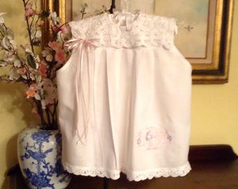 May include: White sleeveless dress with a lace collar and a pink ribbon. The dress has a pleated skirt and a small embroidered design on the front.
