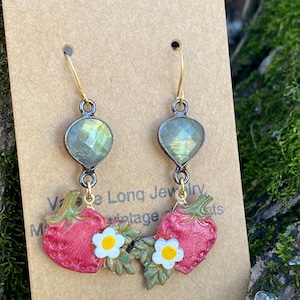 May include: A pair of dangle earrings featuring a green gemstone and a red strawberry charm with white flowers. The earrings are made with gold-tone metal.