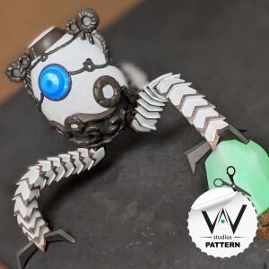 Puede incluir: Un robot blanco y gris con un ojo azul, brazos articulados y un diseño detallado. El robot está posado sobre una superficie verde con un par de tijeras.