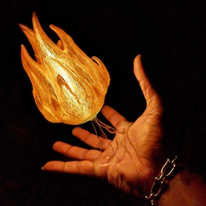 May include: A hand holds a glowing, golden, flame-shaped object with thin, wispy tendrils.