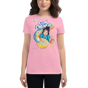 May include: A light pink t-shirt featuring a cartoon cereal box design. The design includes the text "CON' CRUNCH" above a bowl of cereal with a cartoon character. The shirt is made of a soft material.