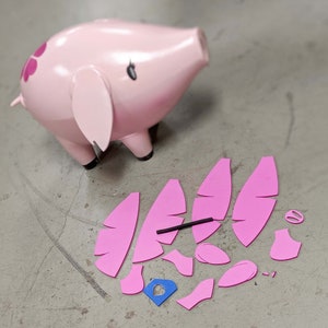 May include: A pink pig figurine with black accents and a floral design on its side. Several pink foam pieces are scattered nearby, including ear and wing shapes, suggesting a craft or DIY project.