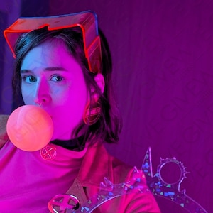 May include: A person wearing a pink shirt and a brown jacket with a large, clear, plastic, geometric design. The person is wearing a red and clear plastic visor and has a large, pink bubble in their mouth.