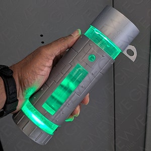 May include: A gray cylindrical container with a green glowing liquid inside. The container has a textured surface and a small loop on the top.