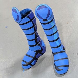 May include: A pair of blue boots with black geometric designs. The boots have a high shaft and a rounded toe. The design includes stripes and curved lines, with a blue and black color scheme. The boots are on a gray surface.