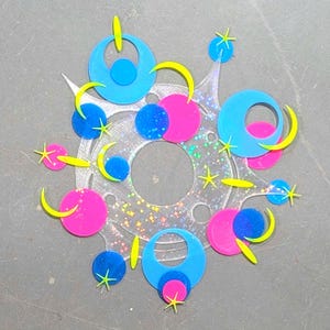 May include: A circular, clear acrylic ornament featuring a holographic star pattern. It is decorated with layered, colorful acrylic shapes including bright blue rings, solid pink and blue circles, and small yellow crescent moons and star accents.