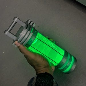 May include: A cylindrical container with a glowing green liquid inside. The container has a metallic frame and handle. The liquid appears to have bubbles. The container is held by a person.