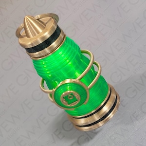 May include: A cylindrical object featuring a translucent bright green body with horizontal ridges, accented by metallic gold-colored rings and a pointed top cap. A circular gold-colored emblem with a square center is attached to the side.