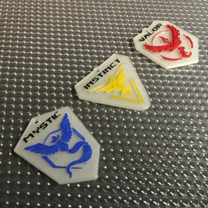 May include: Three translucent, geometric pendants with team logos. The pendants are labeled "Mystic" with a blue bird, "Instinct" with a yellow bird, and "Valor" with a red bird. The pendants are on a textured, silver surface.
