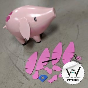 May include: A pink pig-shaped bank with black hooves and a small black eye. Pink paper cutouts of wings and other shapes are next to the bank. A pair of scissors and a blue cutout are also present. The bank has a glossy finish.