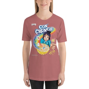 May include: A pink t-shirt with a cartoon illustration of a person holding a bowl of cereal. The cereal box says "Con' Crunch" and "Part of the Crunch Family".