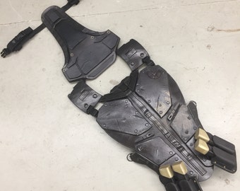 May include: A black and gray tactical vest with straps and gold accents. The vest has a textured, segmented design and appears to be made of a durable material.
