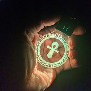 May include: A glowing ankh-shaped keychain. The ankh symbol is in the center of a clear, circular pendant, surrounded by hieroglyphic-style symbols. The keychain is held in a hand against a dark background, with a bright green glow.