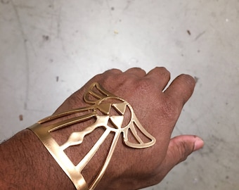 May include: A gold-colored metal bracelet with a detailed design featuring a stylized winged symbol. The bracelet is worn on a person's wrist.