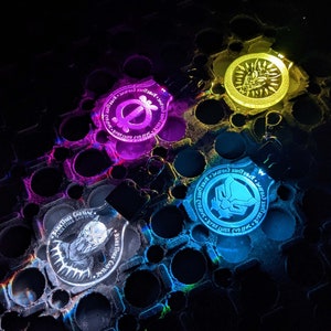 May include: Four glowing keychains with different designs and colors. One keychain glows purple, another yellow, another blue, and the last one white. Each keychain has a different design, including a symbol, a face, and a panther.