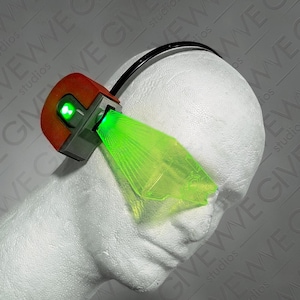 May include: A futuristic, orange and gray device with a green laser beam projecting from it. The device is attached to a black headband and is mounted on a white head form. The laser beam is a bright green color.