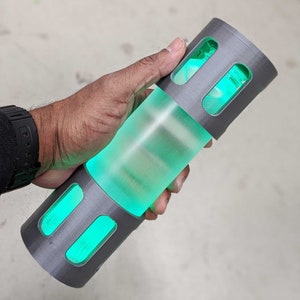 May include: A gray and green cylindrical container with a translucent body and a textured gray cap. The container has three rectangular openings that reveal the green liquid inside.