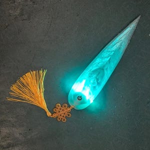 Pode incluir: Uma luz em forma de folha, de cor turquesa, com uma borla e um nó decorativo. A luz tem um botão de ligar/desligar e emite um brilho brilhante e etéreo. A borla é de cor laranja dourado.