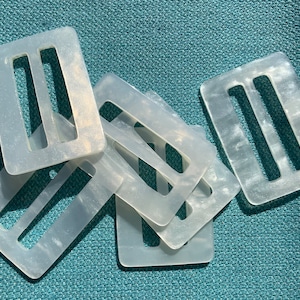 May include: A collection of rectangular, off-white plastic belt buckles with three slots. The buckles have a pearlescent, marbled appearance and are scattered on a teal fabric surface. These are suitable for various crafting and sewing projects.