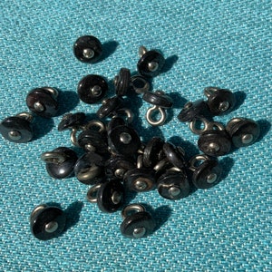 May include: A collection of small, black buttons with a shiny, domed surface and a silver-colored metal loop on the back. The buttons are scattered on a textured, teal-colored surface, creating a contrast between the dark buttons and the light background.